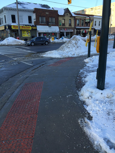Street corned in winter scene.  Clear pavement with snow piles around.  White markings for crosswalk on street and truckated domes in sidewalk edge. 