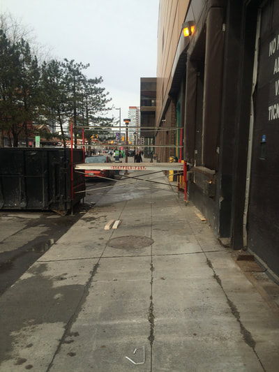 View down sidewalk with temporary walkway blocking path between building and dumpster. 