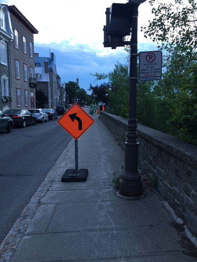 Orange construction side with left facing arrow positioned on narrow sidewalk. 