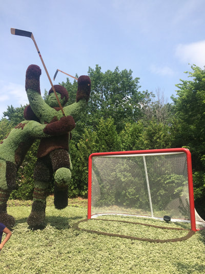 Statue of hockey player made of grass stands to left of a large hockey net. 